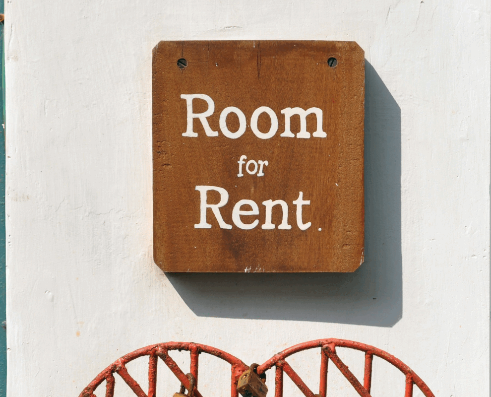 Wooden “Room for Rent” sign on white wall with teal framing and red decoration, symbolising the softening rental market